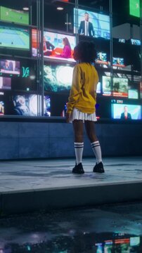 Vertical Screen: African Woman in a Yellow Jacket Stops to Watch Multiple News Channels on a Storefront TV Display at Night. Neon Screens Reflecting on a Wet Pavement as Anchor Reporting News