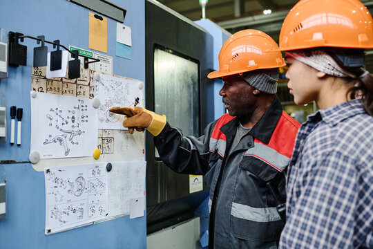 Two factory workers examining detailed blueprints pinned on a corkboard discussing important aspects of the technical drawings in an industrial setting