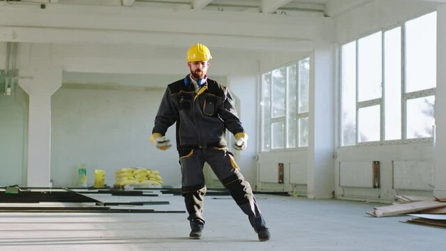 Attractive young engineer in a safety helmet and uniform dancing excited and happy at the end of the work day at modern construction building