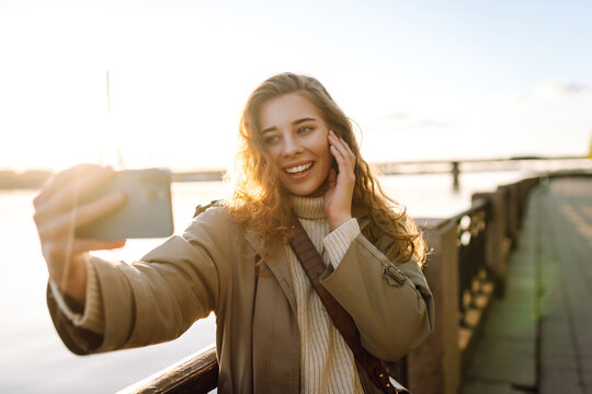 Selfie time. Young woman holding mobile phone taking selfie photo using smartphone camera standing on waterfront. Lifestyle, travel, tourism, vacation, technology, weekend.