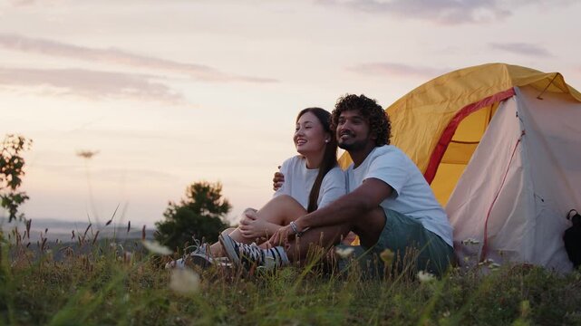 Young couple of Indian guy and Asian girl enjoying the summer's evening  and sunset sitting near the tent in the mountains.