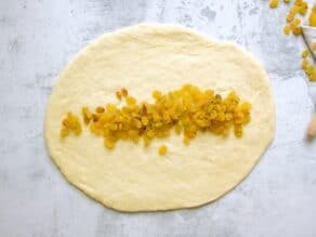 Overhead shot of challah dough that has been rolled into an oval shape. Golden raisins are spread horizontally across the center of the dough.