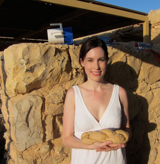 A photo of Tori Avey on top of the historical fortress Masada, holding braided challah dough.