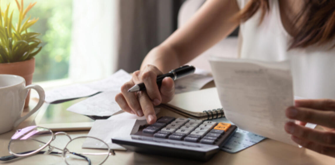 a calculator sits on a table, a hand holding a pen is using the calculator