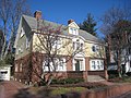 A two story Colonial Revival house. The first floor is brick with bay windows on either side of the front door, the second floor is painted yellow wood siding. Brick walls flank the sidewalk leading up some steps to the front door landing.