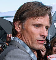 A profile photograph of Viggo Mortensen, a light-haired man wearing a striped collared shirt and a grey blazer.