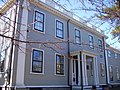 A gray two story house with white trim surrounding black-framed windows. The front door is sheltered by a small pillared porch, and there is a balustered railing around the roof.