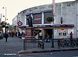 A grey-bricked building with a rectangular, dark blue sign reading "TOOTING BROADWAY STATION" in white letters all under a grey sky