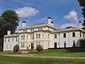 An expansive mansion: it has a large roughly square central section that is three stories with two story wings extending from either side. Square window boxes are built out from the central section. The house is light blue with white trim.