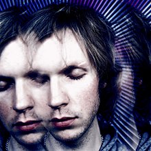Retrato en primer plano del músico Beck con los ojos cerrados sobre un fondo azul abstracto.