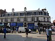 A white building with a blue-shingled roof and a blue sign reading "FINCHLEY ROAD STATION" in white letters all under a blue sky with white clouds