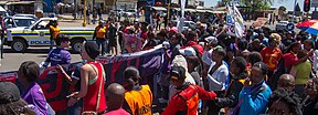 A parade of people walking holding banners and signs. A people car is in the background