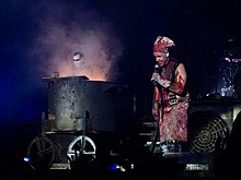 Lindemann blutverschmiert mit Kochmütze und Schürze neben einem dampfenden Kochtopf, in dem Lorenz mit weiß geschminktem Gesicht und Sonnenbrille Keyboard spielt.