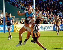 Two young male athletes contest the football. One is wearing a light blue, black and white sleeveless shirt and white shorts, while the other is wearing a brown and gold striped sleeveless shirt and brown shorts.