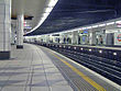 The interior of a building with a railway track running between white-tiled pillars upholding a white-and-black ceiling lit by electrical lights