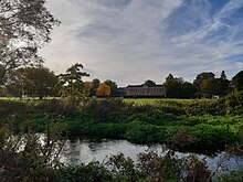 Photograph of a river in front of wide playing fields and distant buildings