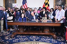 Governor Moore signing a proclamation recognizing International Transgender Day of Visibility, surrounded by trans rights advocates and seated alongside Susan C. Lee and Aruna Miller.