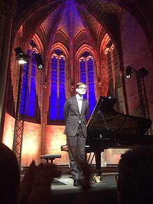 Víkingur Ólafsson wearing a dark suit, standing in front of a grand piano inside a church, appearing to bow slightly with hands behind his back