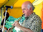 A man wearing a patterned dress shirt, performing a wind instrument while standing behind a microphone