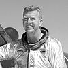 A smiling and energetic blonde man wearing a pressure suit stands in front of an airplane