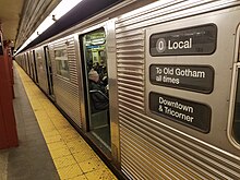 A corrugated silver metal subway train sits with its doors open in a station. Its rollsign reads "0 Local / To Old Gotham all times / Downtown & Tricorner".