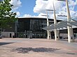 A grey building with grey lampposts in front and a green tree in the foreground all under a light blue sky with billowy, white clouds