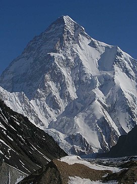 A large angular white mountain, with steeply sloped sides mostly covered with snow, dominates surrounding brown mountains