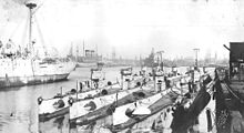 Nine submarines sit moored next to each other against a dock, with ships in the background