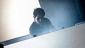 Porter Robinson stands at a table with a brightly-lit screen; another screen is behind him