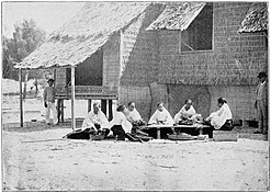 A group of women seated around a table with traditional houses in the background