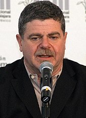 A 56-year-old man with dark gray hair talking into a microphone, looking at something beneath the camera.