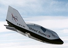 A small, stubby spaceplane, coloured black on its underside and white on its topside, descending against a cloudy sky. The words "United States" and the NASA logo are visible on its sides.