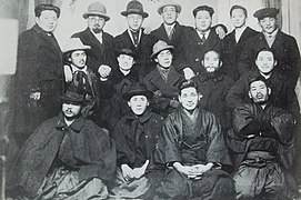 Photo d'un groupe d'hommes sur trois rangées. Ils portent des costumes traditionnels du début du XXe au Japon, ainsi que des chapeaux de ville.