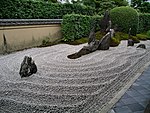 Rock garden with raked gravel and large standing stones in front of a small wall and green plants.