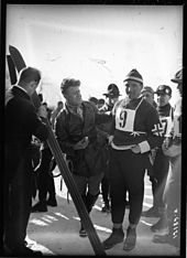 Photographie en noir et blanc d'un homme portant une tenue sportive avec le dossard numéro 9 et un bonnet, entouré d'autres hommes dont un face à lui tenant une paire de ski.