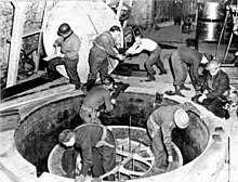 Soldiers and workmen, some wearing steel helmet, clamber over what looks like a giant manhole.