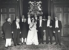 A number of older men in formal dress surround a young woman with a white gown.