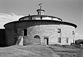 This black and white photo shows a round barn made of fieldstone. It is topped by a white wood frame multi-sided section that is smaller, and then a small turret.