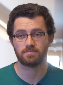 Austin Wintory speaking on video in 2019. He is standing against a blurry office background, wearing a green shirt and glasses