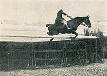 Photo en noir et blanc d'un homme franchissant un obstacle sombre surmonté d'une barre blanche; la position du cavalier est notable car il est très assis dans sa selle, les bras tendus et les rênes longues pour permettre au cheval de sauter.