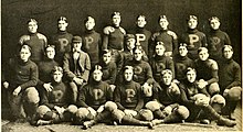 A sepia-tone picture of around thirty young white men, all in American football uniforms from the beginning of the 1900s. They are taking a team picture, staged in rows and all facing directly into the camera. Some are sitting cross-legged.