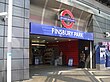 A grey-panelled building with a blue sign reading "FINSBURY PARK" in white letters and a woman with a brown coat standing in the doorway