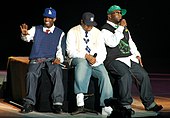 Three African-American men sitting next to each other on a black stage. They are all wearing caps with jeans and sneakers.