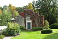 A stepped stone facade frames a green door, above which there is a small round window with white trim. Much of the stonework is covered in ivy, and the building is set in a well manicured garden.