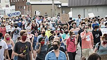 A crowd, facing the camera, is marching along a street, some holding signs like "PROTECT BLACK BODIES", "NO MORE", and "JUSTICE FOR GEORGE".