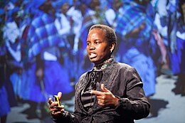 Photograph of an African woman in a grey brocaded suit standing in front of a backdrop depicting a group of African women in blue dresses, while giving a speech. She is wearing a geometrically patterned beaded neck collar, has a yellow device in her right hand, and is pointing with her left index finger to the left of the stage.