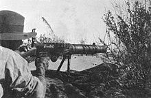 A soldier is looking through the sights of a machine gun amongst the grass in the prone position.