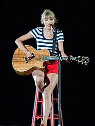 Swift in a white and black striped shirt and red shorts playing an acoustic guitar