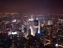An aerial view of skyscrapers at night. Most of the buildings are lit up with lights coming from the inside of them. In the background, a dark sky with no clouds can be seen.