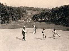 Photo d'un parcours de golf. Au premier plan, un groupe d'hommes est en train de jouer ; au loin, on distingue des forêts.